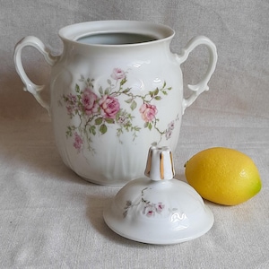 May include: A white porcelain sugar bowl with two handles and a matching lid, decorated with pink rose floral patterns and green leaves. The lid features a gold-trimmed finial. A single yellow lemon is positioned next to the sugar bowl.