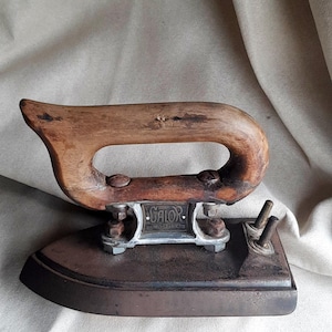 May include: An antique iron with a wooden handle and a metal base. The handle is curved and attached to the iron with metal hardware. The base is rectangular with a pointed front. The iron has the word "GALOR" on it.