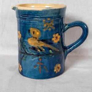 Puede incluir: Una jarra cilíndrica de cerámica con un esmalte azul profundo brillante, decorada con motivos pintados a mano de pájaros y flores amarillas. El interior es de color crema y el recipiente incluye un asa curva y un pequeño pico vertedor.