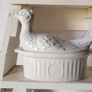 May include: A white ceramic duck-shaped casserole dish with a lid. The duck has detailed feathering and a long neck. The dish is oval with vertical ridges and a circular indentation on the front. The casserole dish is sitting on a white shelf.