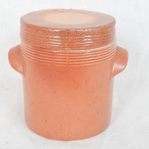 Peut inclure: Un pot cylindrique en céramique de couleur terre cuite avec un couvercle assorti. Le pot présente deux petites anses latérales, une texture mouchetée et des lignes horizontales rainurées entourant la partie supérieure du corps.
