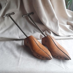 May include: Two vintage wooden shoe stretchers with metal handles and threaded rods. The shoe forms are light brown with a curved shape, and the metal components are silver. The stretchers are designed to maintain the shape of shoes.