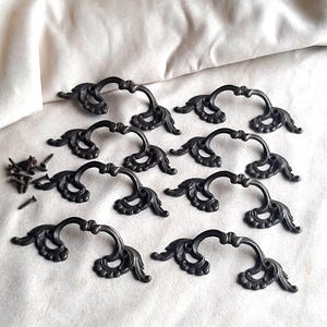 May include: A collection of ten antique-style, black metal drawer pulls with ornate, curved designs. Each pull features a decorative flourish at each end and a curved handle. Several screws are also visible, suggesting a hardware set.