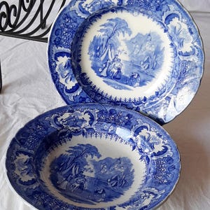 Może przedstawiać: Dwa antyczne, niebiesko-białe ceramiczne talerze. Talerze mają szczegółową scenę krajobrazu na środku, otoczoną ozdobną obwódką. Talerze są prawdopodobnie zabytkowe i dodają odrobinę klasycznej elegancji do każdego nakrycia stołu.