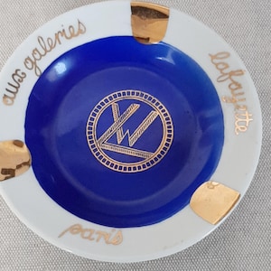 Puede incluir: Un cenicero redondo blanco con un centro azul profundo y detalles dorados. El cenicero presenta las palabras "aux galeries lafayette paris" en escritura dorada alrededor del borde y un logotipo dorado en el centro.