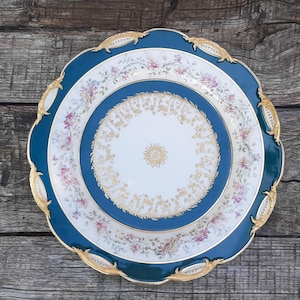 May include: A round porcelain plate featuring a deep teal border with gold-toned ornate accents along the scalloped edge. The centre displays a white circular field with a gold floral motif, surrounded by a ring of delicate pink and purple flowers.