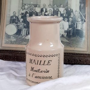 Puede incluir: Un tarro de mostaza Maille rosa claro vintage con texto negro y borde decorativo. El tarro está frente a una fotografía en blanco y negro enmarcada de un grupo de personas. El tarro está sobre una tela blanca.