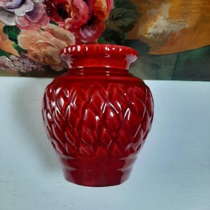 Peut inclure: Un vase en céramique rouge brillant avec un motif en losanges texturé. Le vase a un bord évasé et un corps arrondi. L'arrière-plan présente une peinture florale avec des roses dans des tons de rose, de rouge et d'orange.