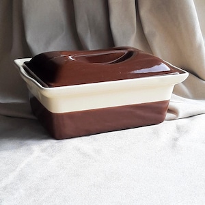 May include: A two-tone ceramic casserole dish with a lid. The top and bottom sections are a deep brown, with a cream-coloured band in the middle. The lid features a recessed handle. The dish is set against a neutral backdrop.