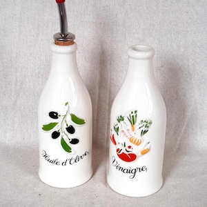 May include: Two white ceramic oil and vinegar bottles. One has a red stopper and the words "Huile d'Olive" with a green olive branch and black olives. The other has "Vinaigre" with a colourful vegetable design.