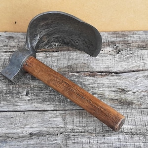 Op de afbeelding: Een handgereedschap met een gebogen, halfrond metalen blad met een donkere, verweerde afwerking, bevestigd aan een cilindrische houten handgreep. Het blad heeft een getextureerd, gesmeed uiterlijk en de handgreep toont een natuurlijke houtnerf.