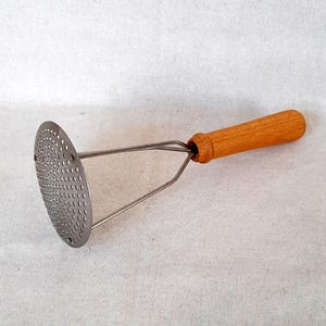 Puede incluir: Un pasapurés con una cabeza perforada de acero inoxidable y un mango de madera. El pasapurés tiene una cabeza circular con numerosos agujeros pequeños y dos brazos de soporte metálicos. El mango es de color marrón claro.