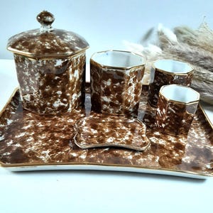 Op de afbeelding: Een vintage toilettafelset met een bruin en wit gespikkeld patroon. De set bevat een pot met deksel, drie kleine kopjes, een zeepbakje en een rechthoekig dienblad met gouden rand. De stukken zijn op een wit oppervlak geplaatst.