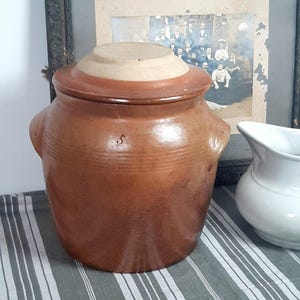 May include: A brown stoneware crock with a lid, featuring a light tan top and a darker brown band. The crock has two small handles and a subtle 'S' marking. It sits on a striped gray and white fabric, with a framed vintage photograph in the background.