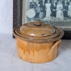 Peut inclure: Un petit pot rond en céramique avec couvercle, glaçure beige et marron. Le pot a deux poignées et un bouton sur le couvercle. L'arrière-plan comprend une photographie encadrée en noir et blanc.
