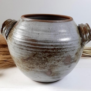 Op de afbeelding: Een handgemaakte keramische pot met een brede, afgeronde body en twee zijhandvatten. De pot heeft een lichtgrijze glazuur met bruine accenten en een donkerbruine rand. De rustieke pottenbakkerij is geschikt voor woondecoratie.