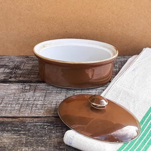 May include: An oval-shaped ceramic casserole dish with a glossy brown exterior and a smooth white interior. The dish includes a matching brown lid with a small central handle, featuring a clean rim and a sturdy, deep base for food preparation.