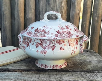 Op de afbeelding: Een witte keramische terrine met deksel, voorzien van een donkerrood bloemen- en rankenpatroon. De terrine heeft twee zijhandgrepen en een bijpassende handgreep op het deksel, rustend op een ronde voetstuk.