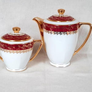 Peut inclure: Service à thé en porcelaine blanche avec bordure dorée, comprenant une théière et un sucrier. L'ensemble présente une bande rouge avec des motifs de filigrane dorés. La théière a une anse et un bec courbés.
