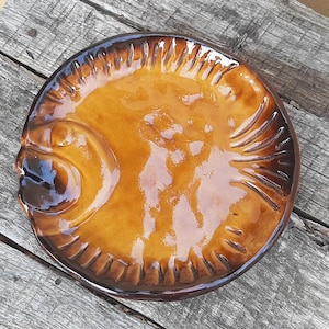 Könnte beinhalten: Ein runder Keramikteller mit glänzender bernsteinfarbener und brauner Glasur, der eine in das Design integrierte Fischform aufweist. Der Rand ist mit strukturierten, strahlenförmigen Vertiefungen verziert, die an Flossen erinnern.