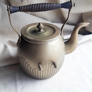 May include: Antique brass tea kettle with a dark wooden handle and a decorative spout. The kettle has a round body with vertical ribbed detailing and two small knobs on the front. The lid is topped with a small, decorative finial.