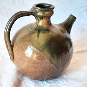 May include: A handcrafted ceramic pitcher with a rounded body and a spout. The pitcher features a handle and is glazed in shades of brown and green. The pottery piece is likely used for pouring liquids.