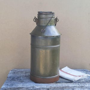 May include: A tall, cylindrical metal milk can with a weathered, muted olive-green finish. It features a narrow neck, a secure lid with a small attached chain, and two side handles. The base is a distinct, darker brown band, showing signs of age.