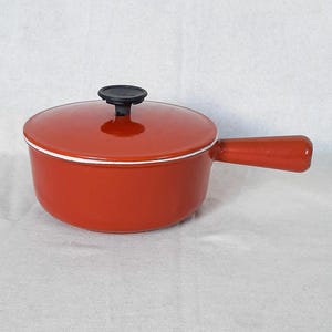 May include: A small, red enameled cast iron saucepan with a lid and a single handle. The lid has a black knob. A white stripe accents the rim of the pan. The pan is designed for stovetop cooking.