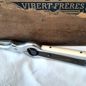 Könnte beinhalten: Vintage Nussknacker aus Metall mit silbernem Finish und hellem Holzgriff. Das Werkzeug hat ein spitzes Ende und ein Scharnierdesign zum Knacken von Nüssen. Der Hintergrund zeigt eine Holzoberfläche mit dem Text "VIBERT FRERES".