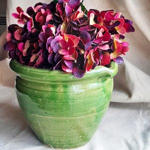 May include: A vibrant green ceramic pot filled with a lush arrangement of deep purple and red hydrangeas. The pot has a rounded shape with horizontal ridges and a glossy finish, creating a striking contrast with the flowers.