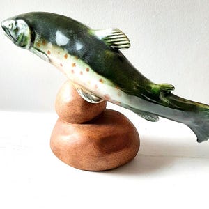 Könnte beinhalten: Eine Keramikfischskulptur mit grün-weißem Körper, gesprenkelt mit orangefarbenen Punkten, ist in einer Sprungpose dargestellt. Der Fisch ruht auf einem braunen, abgerundeten Sockel. Die Skulptur steht vor einem weißen Hintergrund.