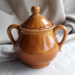 Op de afbeelding: Een bruine keramische suikerpot met een deksel en twee handvatten. Het deksel heeft een kleine knop en een bladeren ontwerp. De kom heeft een glanzende afwerking en is waarschijnlijk vintage. De suikerpot is een keukenartikel.