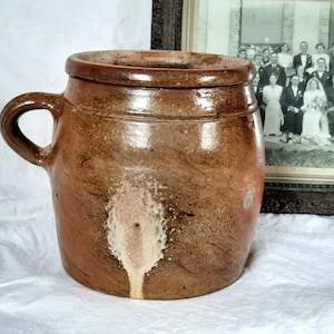 May include: A brown stoneware crock with a handle, featuring a wide opening and a slightly flared rim. The antique jar has a rustic appearance with a light-coloured drip pattern on the front. A vintage photograph is in the background.