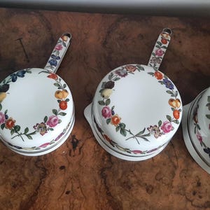 May include: Four white ceramic saucepans with floral designs. Each pan has a handle and a rim decorated with colorful flowers, fruits, and green leaves. The pans are arranged on a wooden surface.