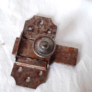 Può includere: Un chiavistello metallico arrugginito con pomello circolare e piastre rettangolari. Il chiavistello presenta un aspetto usurato con segni visibili di età e utilizzo. Il metallo è di colore marrone e argento.
