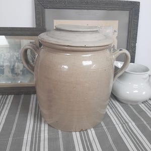 May include: A large, tan stoneware crock with a matching lid and two handles. The antique jar is displayed on a gray and white striped tablecloth. Two framed pictures and a white pitcher are in the background.