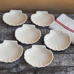 Peut inclure: Un ensemble de six coupelles en céramique blanc cassé en forme de coquilles Saint-Jacques avec des bords cannelés, accompagné d'une serviette en lin beige pliée ornée de deux fines rayures rouges horizontales.