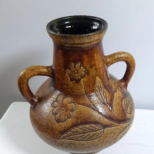 Puede incluir: Un jarrón de cerámica marrón con dos asas y un interior oscuro. El jarrón presenta diseños florales y de hojas en relieve. El exterior tiene un acabado texturizado. Es un objeto decorativo.