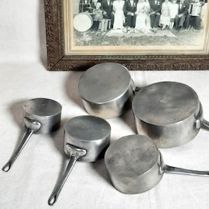 Puede incluir: Un conjunto de cinco cacerolas de metal vintage con mangos largos. Las cacerolas están dispuestas sobre una superficie blanca, con una fotografía en blanco y negro enmarcada en el fondo. La fotografía representa a un grupo de personas.