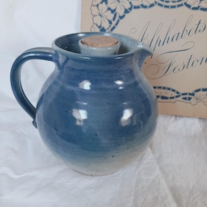 Puede incluir: Una jarra de cerámica azul con asa y boquilla, con base azul más clara. Un inserto de cerámica azul claro más pequeño contiene un tapón de corcho. El fondo incluye un libro vintage con diseños florales y el texto "Alphabets Festons."