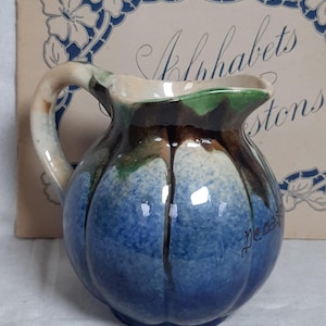 Puede incluir: Una pequeña jarra redonda de cerámica con asa. La jarra es principalmente azul con detalles marrones y verdes. La palabra "decass" está escrita en cursiva en el lateral. El fondo presenta un libro vintage con diseños florales.