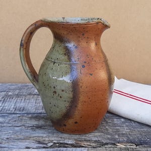 Pode incluir: Um jarro de cerâmica com um esmalte de dois tons, apresentando uma secção verde-sálvia salpicada e uma secção castanho-terracota quente. O recipiente tem um corpo arredondado, uma pega curva no lado esquerdo e um pequeno bico vertedor.