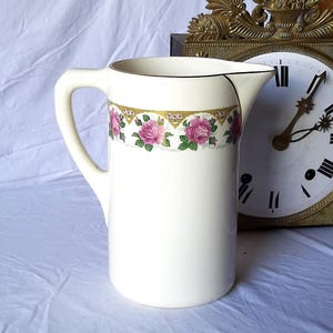 Può includere: Brocca in ceramica color crema con un motivo floreale a rose. La brocca ha un manico curvo e un bordo nero attorno al bordo. Una fascia dorata e floreale decora il centro. Un orologio vintage è sullo sfondo.