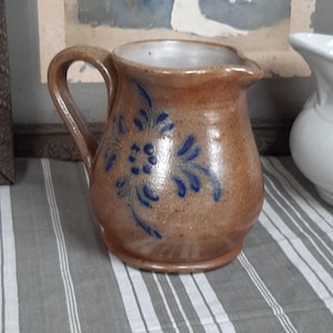 Puede incluir: Una jarra de cerámica marrón con asa y pico vertedor. La jarra está decorada con diseños florales azules. La jarra está sobre un mantel a rayas grises y blancas. Una fotografía enmarcada está en el fondo.