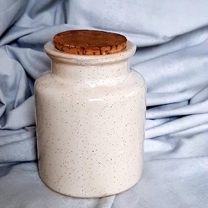 Op de afbeelding: Een gespikkelde, gebroken witte keramische pot met een kurken deksel. De pot heeft een cilindrisch lichaam en een licht uitlopende rand. De kurken deksel is lichtbruin van kleur. De pot staat op een lichtblauwe stof.