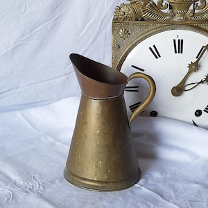 Op de afbeelding: Een vintage messing kan met een koperen rand en handvat. De kan heeft een conische vorm en een tuit. De achtergrond omvat een wit tafelkleed en een goudkleurige klok.