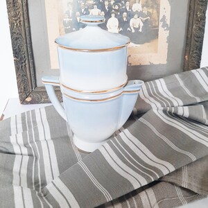 May include: A light blue ceramic coffee server with a lid and gold trim. The server sits on a gray and white striped cloth, with a framed vintage photograph in the background. The server has a handle and a spout.