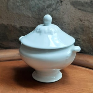 Peut inclure: Un sucrier en céramique blanche avec un couvercle et de petites poignées. Le bol a une base arrondie et un petit bouton sur le dessus du couvercle. Le sucrier est posé sur une surface en bois.