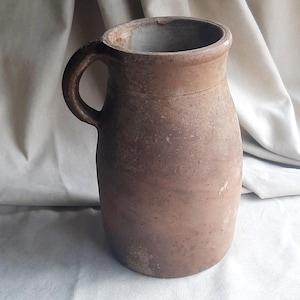 Op de afbeelding: Een bruine keramische kan met een handvat. De kan heeft een cilindrisch lichaam en een brede opening. De buitenkant heeft een getextureerd, verouderd uiterlijk, wat suggereert dat het een antiek of vintage stuk is. De achtergrond is een neutrale stof.