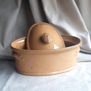 Puede incluir: Una fuente de cerámica ovalada de color beige con tapa. La tapa tiene un pequeño pomo decorativo. La fuente tiene asas a cada lado y un acabado ligeramente brillante. La tapa está parcialmente dentro de la fuente abierta.